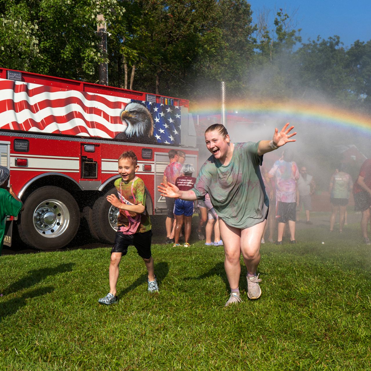 Children run in the rainbow spray of a water hose