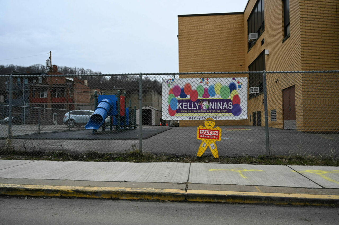 A sign that read's Kelly Ninas, "Where the place is small but the love is big" hangs on a chain-link fence in front of a tan brick building with a small playground behind the fence.