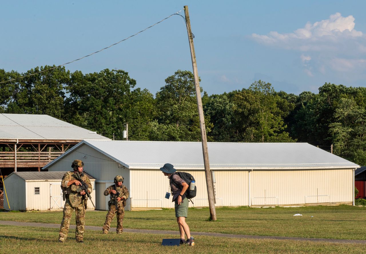 Man in fatigues and gun questions a person wearing shorts