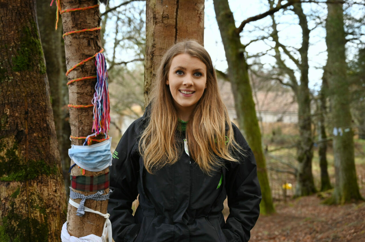 A woman in a black jacket with long blond hair stands next to a thin tree that is wrapped in colorful fabric and masks.