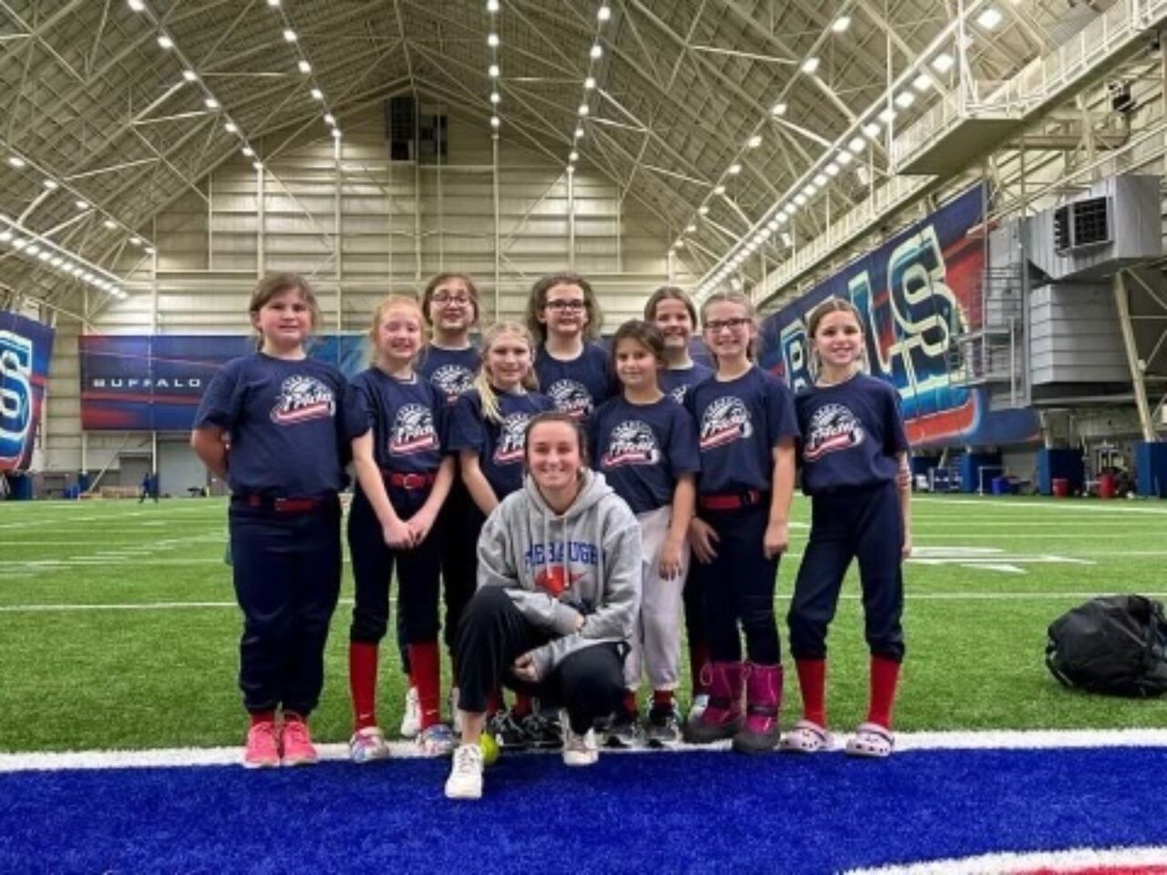 A softball coach kneels on the ground with nine of her softball players behind her. The team is inside a sporting complex with grass in the background.