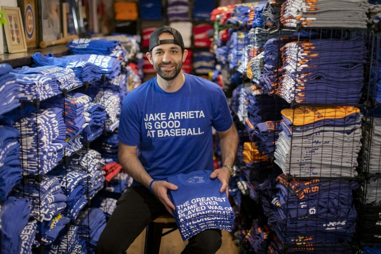 A man with a dark beard sits on a stool wearing a backwards baseball cap and a royal blue tee shirt that reads Jake Arrieta is Good at Baseball in all capital letters. He is surrounded by stacks of tee shirts in black metal racks.