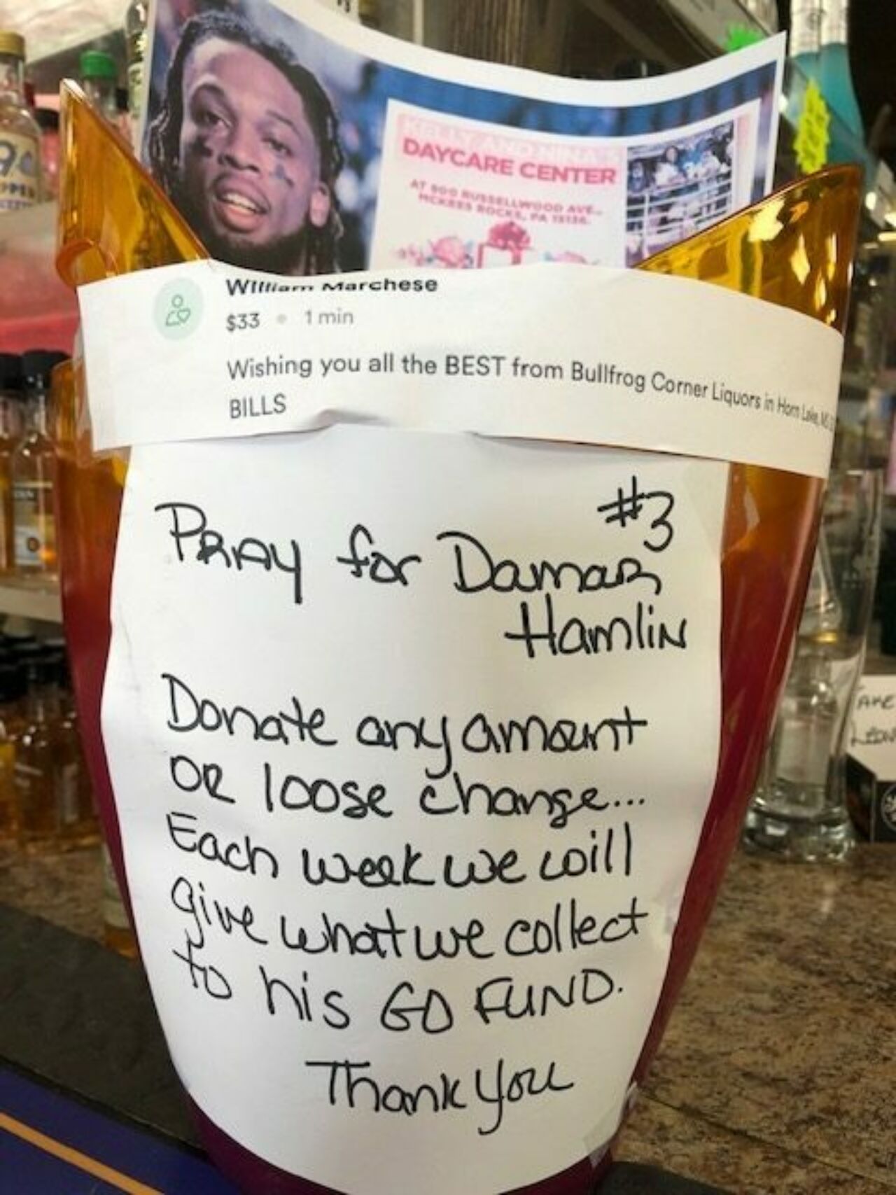 A handwritten note in black on white paper hangs on a donation bin and reads Pray for Damar Hamlin, #3. Donate any amount or loose change. . . each week we will give what we collect to his GO FUND. Thank You.