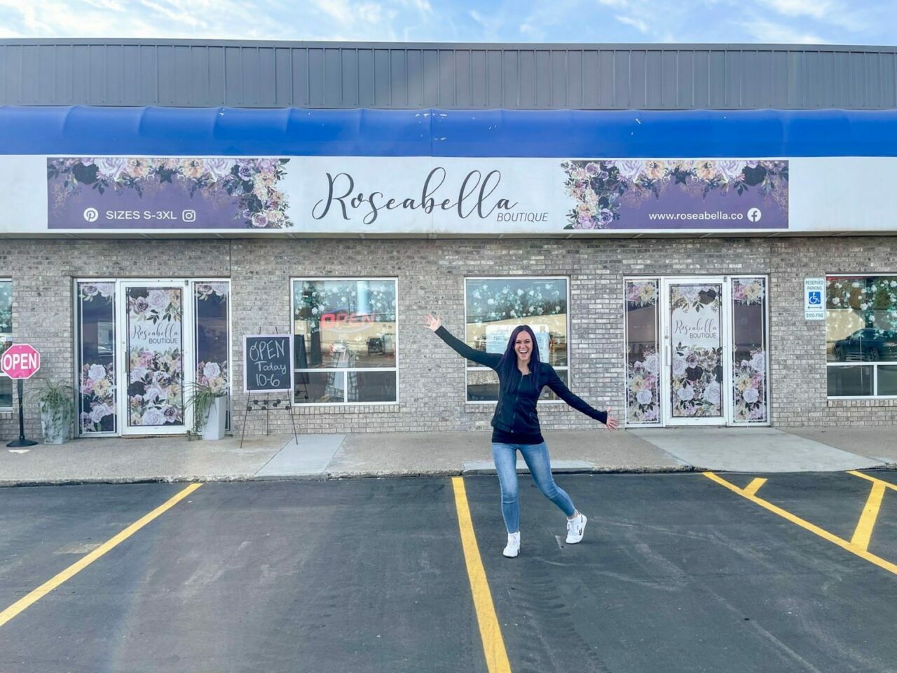 A woman with shoulder-length brown hair poses in front of a storefront signed as Roseabella Boutique in jeans and a black zip up sweatshirt.