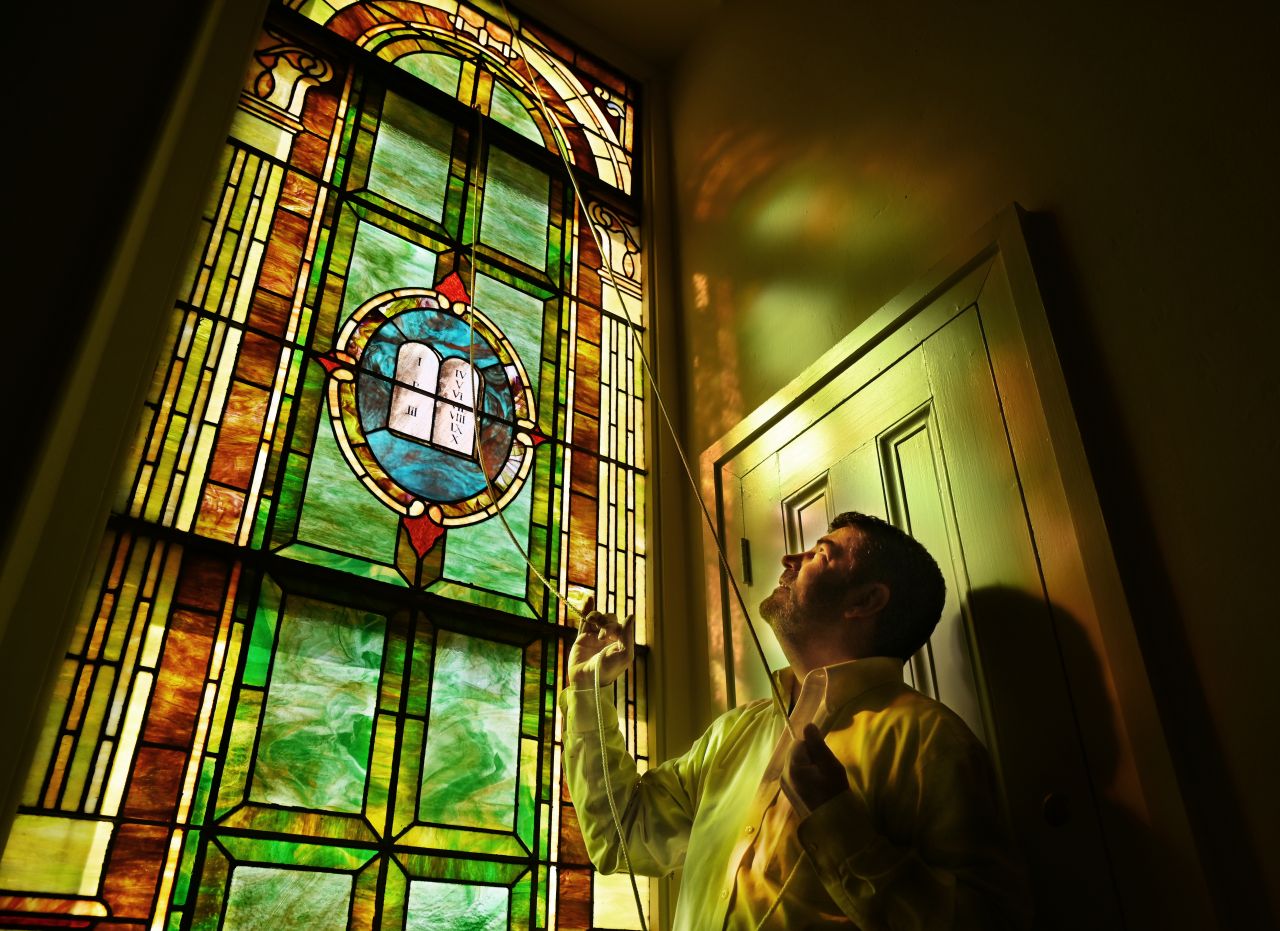 A man in a white collared shirt, reflected in a stained glass window pulls a rope to open the window.