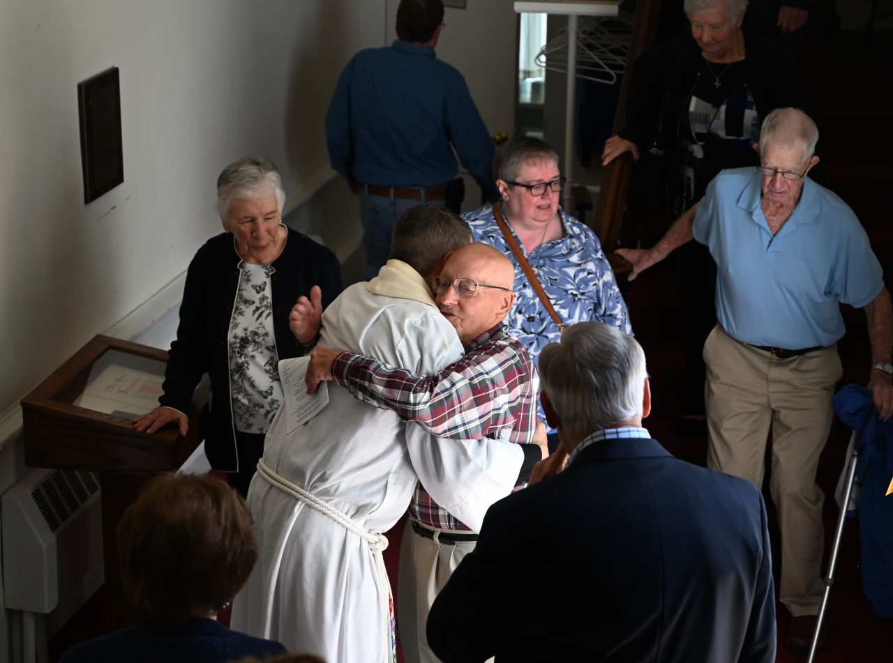 A man in a white robe hugs an older, balding man wearing glasses.