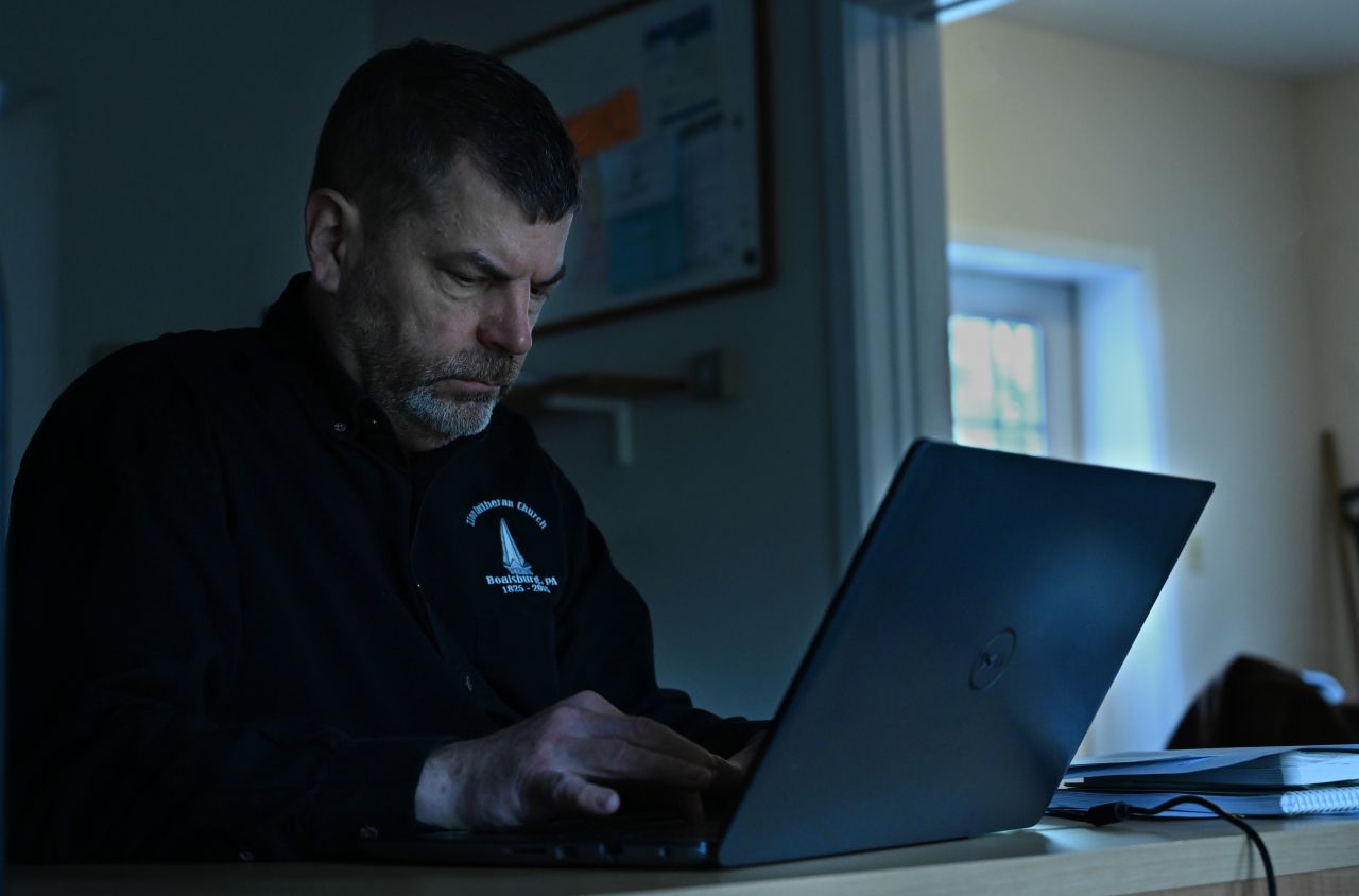 A dark-haired man in a black sweatshirt works at his laptop.