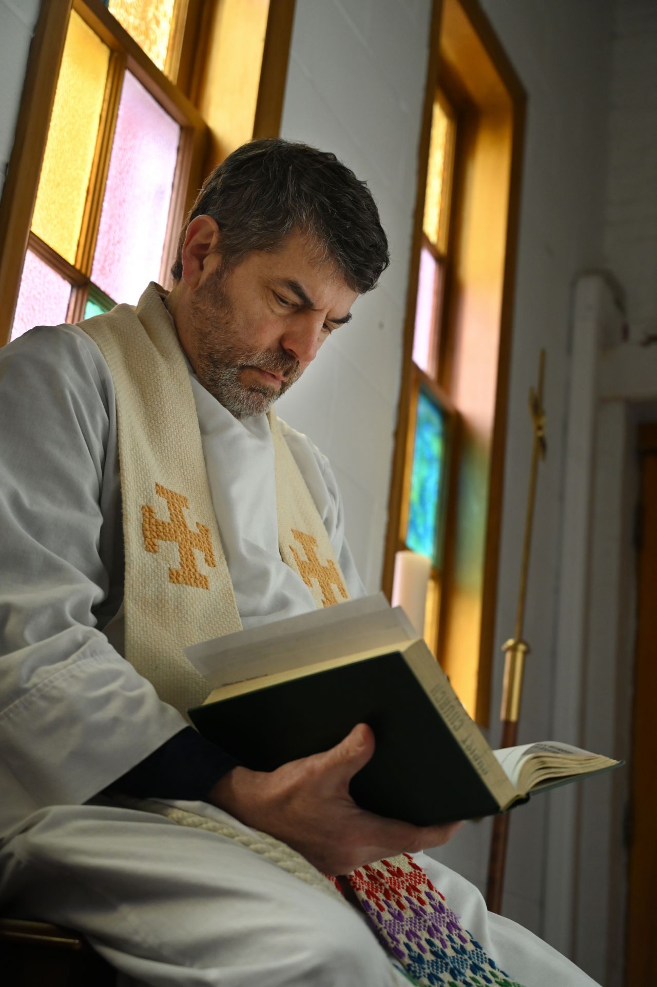 A dark haired man sits alone in front of a stained-glass window and reads the Bible.