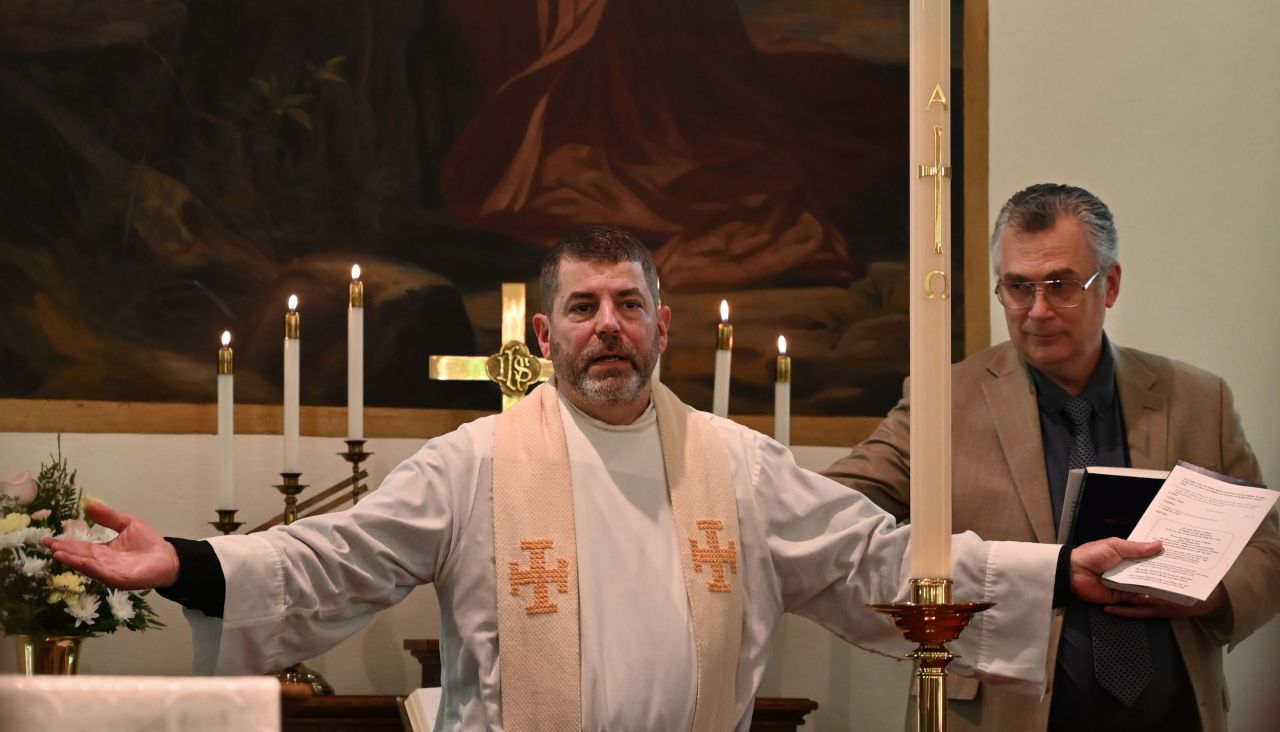 A dark-haired man with a graying beard spreads his arms while delivering a message.