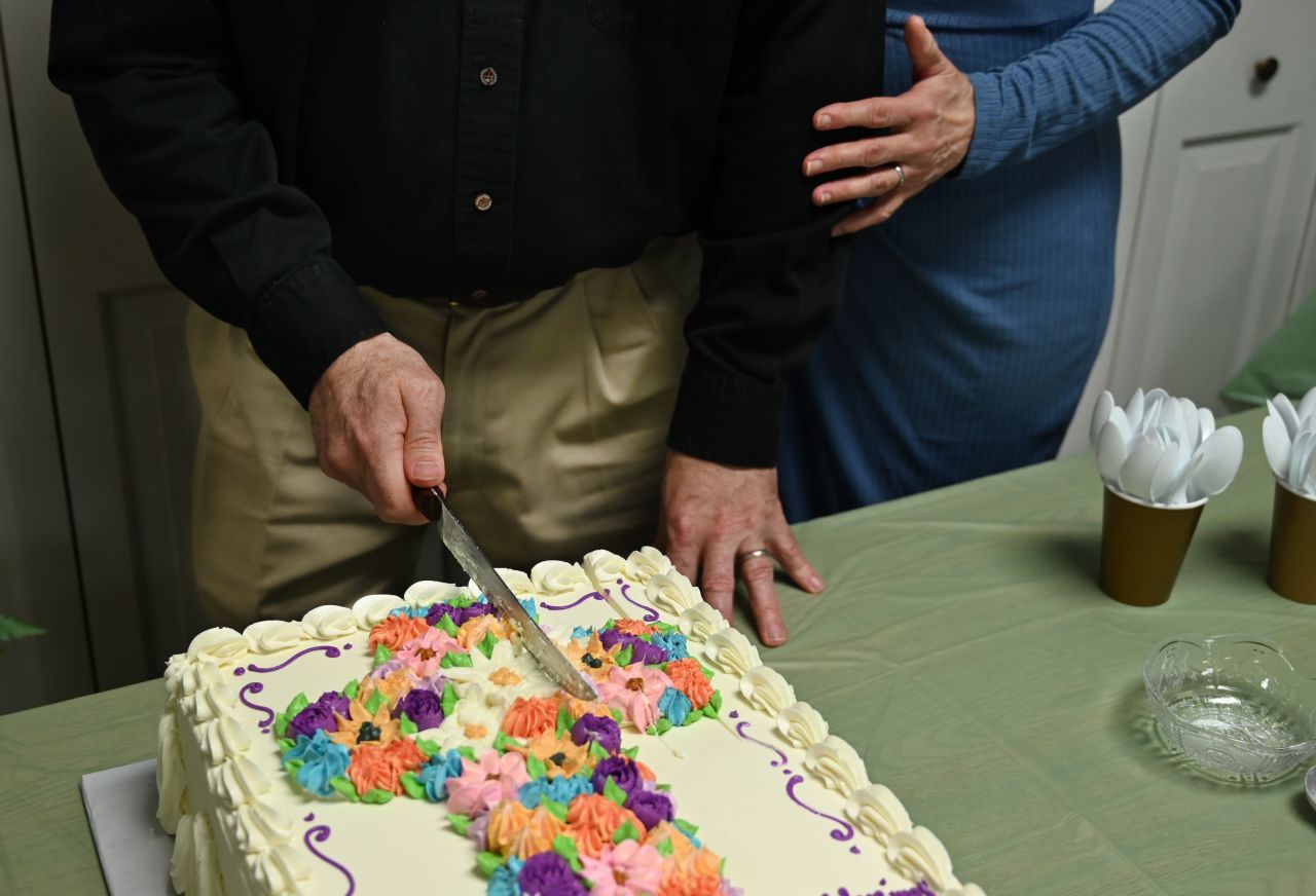 A right-handed man slices through a sheet cake featuring colorful flowers in the shape of a cross.