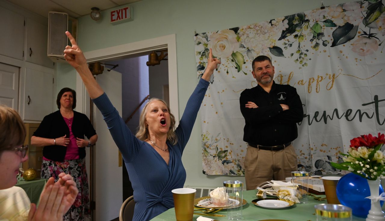 A woman in a blue shirt raises both her ams in celebration while seated at a table.