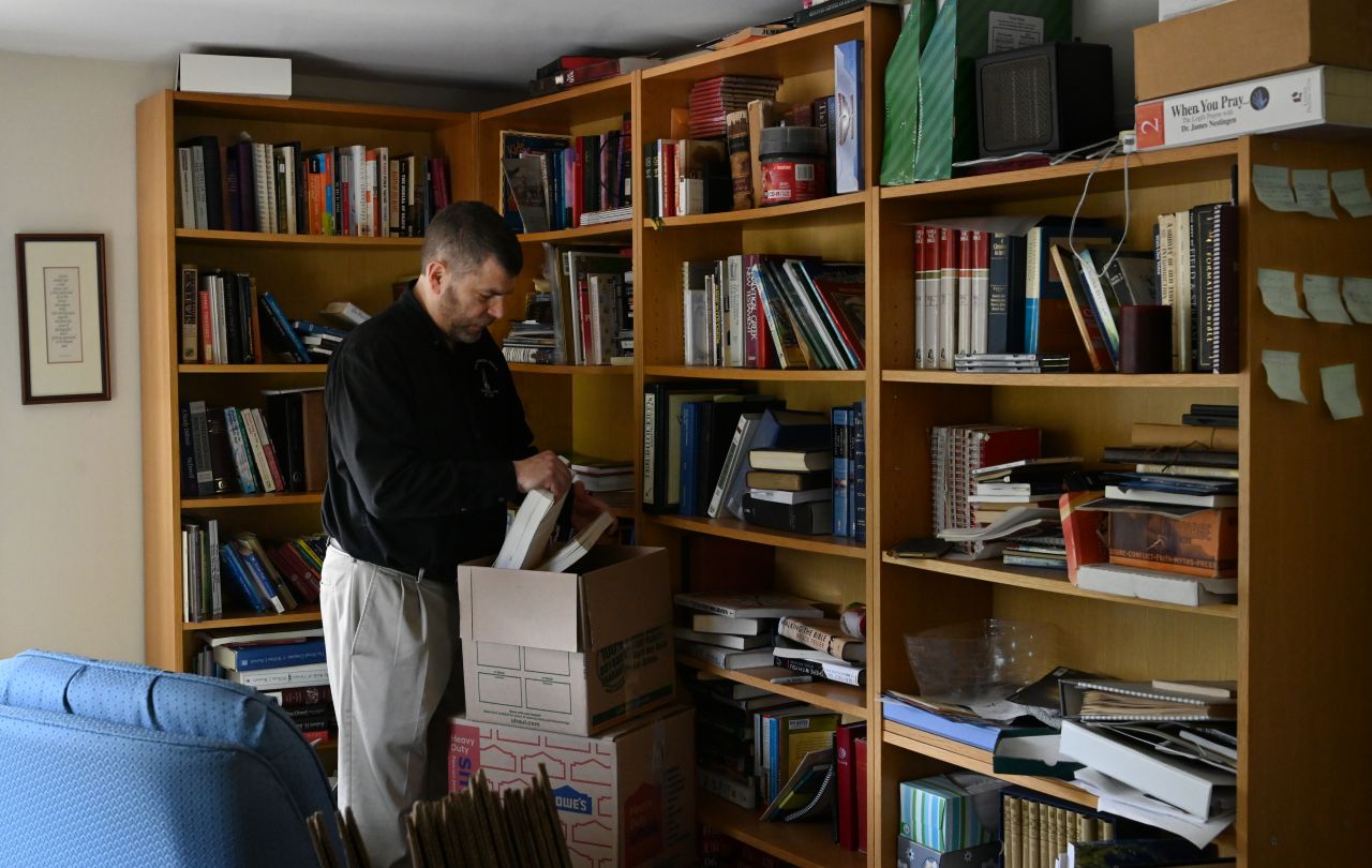 A dark-haired man in a black button-down shirt pulls books from a bookshelf and places them in a box.