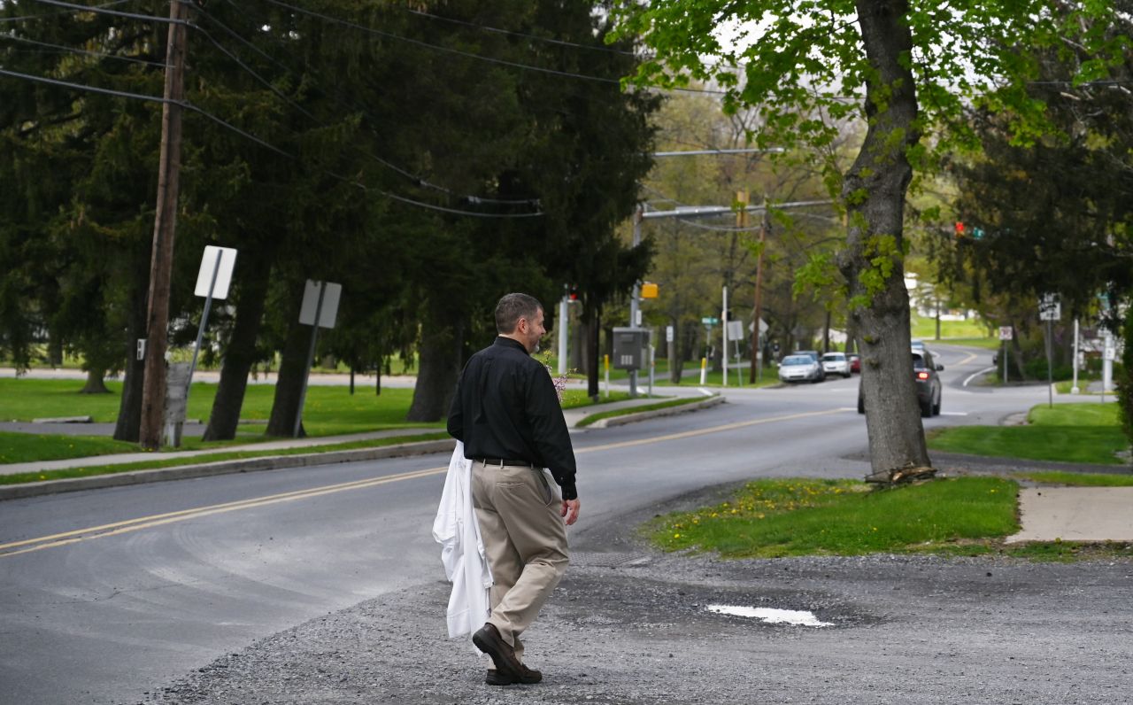 A dark haired man carrying a white robe in his left arm crosses the street.