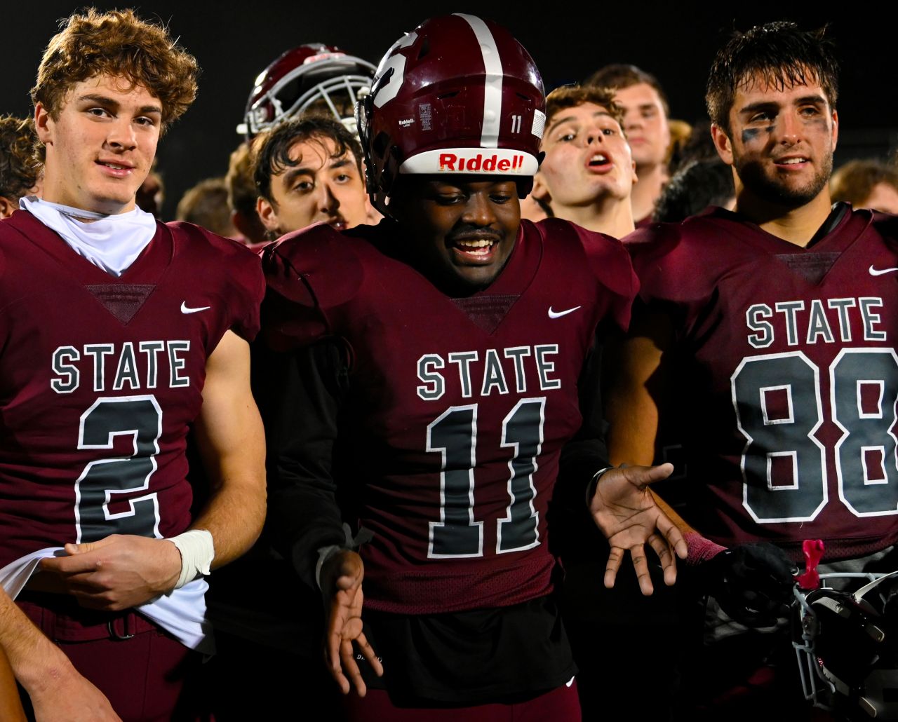 Three high school boys in maroon football jerseys stand in front of their team.