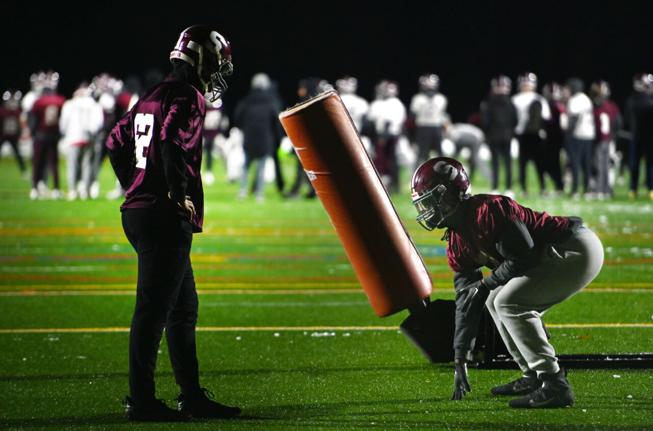 A football player on the left in a maroon jersey, black pants, and a maroon helmet with the capital letter S on the side watches as another player in uniform displays his form for crouching at the line of scrimmage.