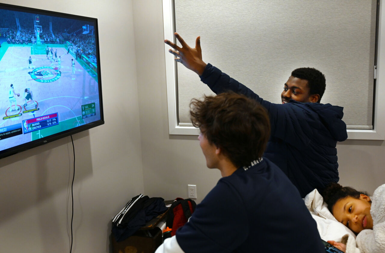 Two teens in navy shirts sit in front of a tv monitor playing a basketball video game.