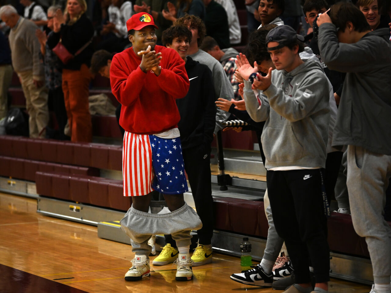 Several teens stand on the side of a wood basketball floor clapping their hands. One is a black teen wearing a red Bass Pro Shop cap, red sweatshirt, and has shorts on with the pattern of the Unites States flag. His friend has a backwards black baseball hat and gray hoodie sweatshirt.