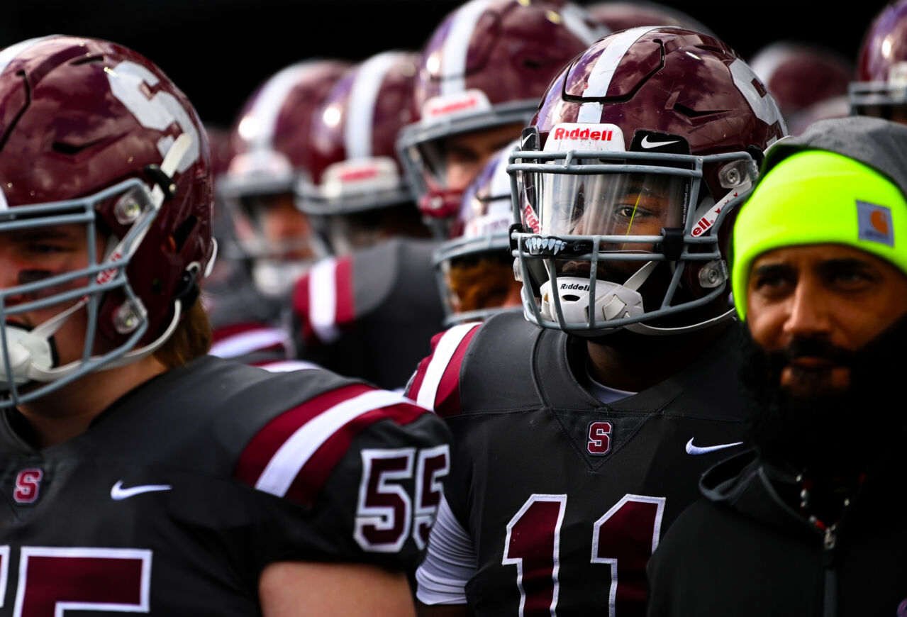 Several members of a football team in dark grey uniforms with maroon helmets emblazoned with a capital S stand awaiting the start of a game. Number 11 is featured in the foreground with a clear plastic face sheild.