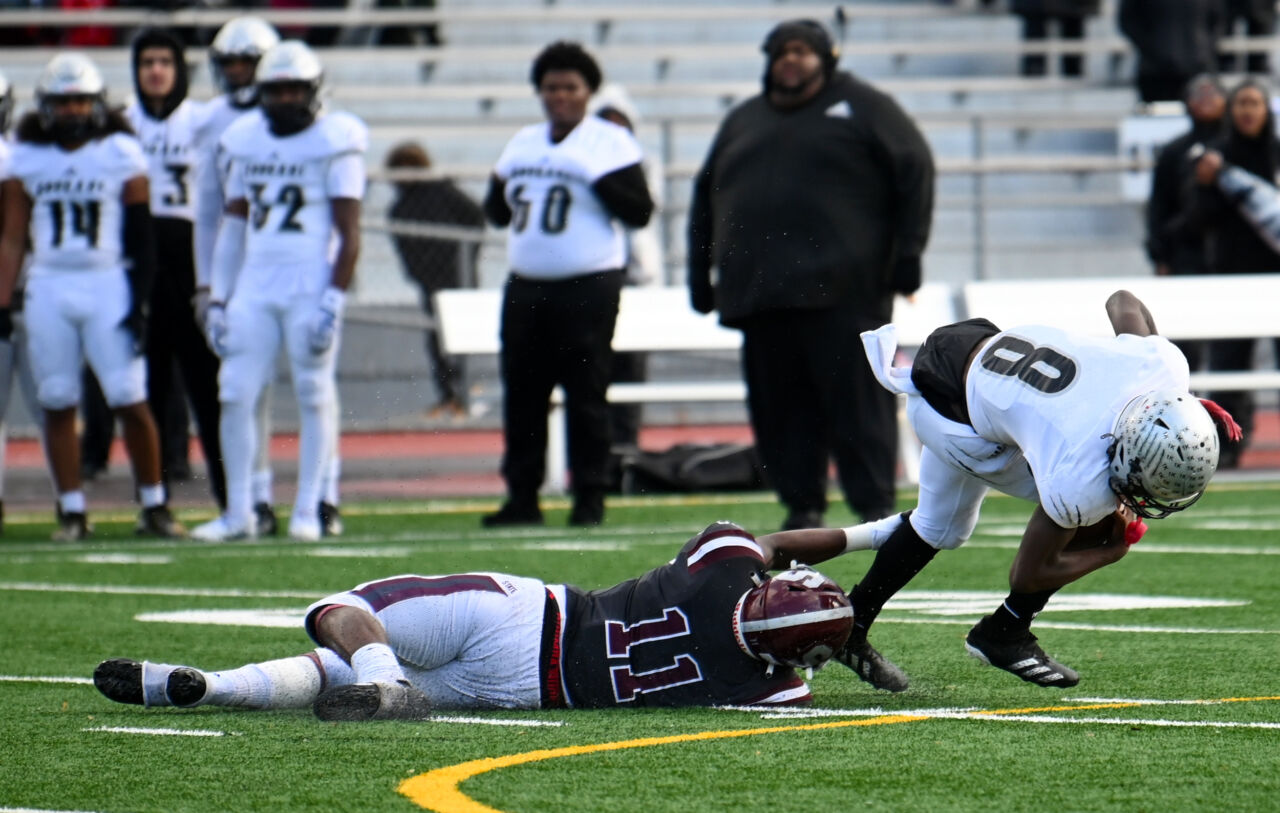 Football player number eleven wearing a dark gray jersey with maroon helmet is laid out on the turn as he grabs the left leg of a quarterback dressed in white uniform and number eight leading to a sack.