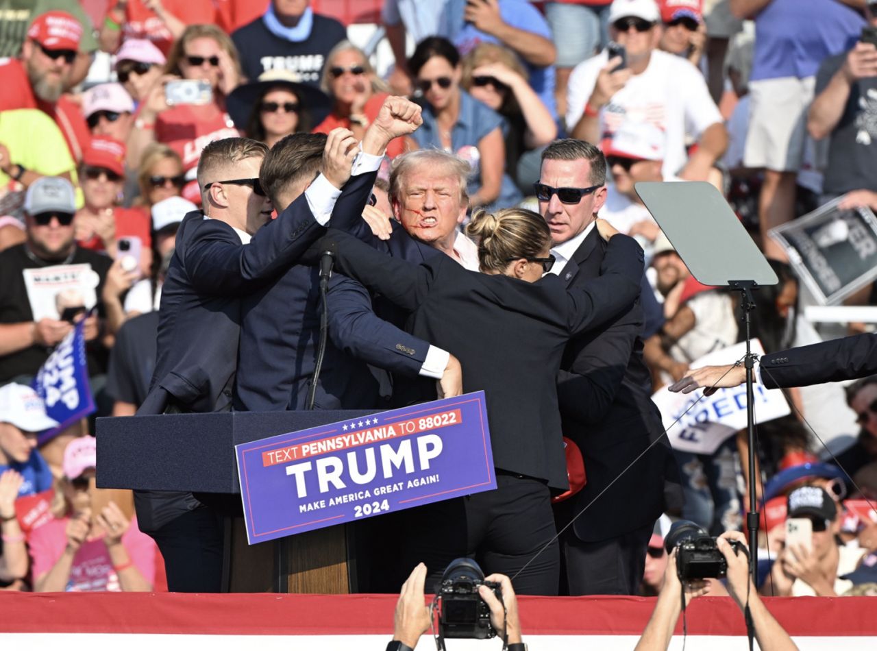 Man with fist raised wearing suit while others in suits crowd around him