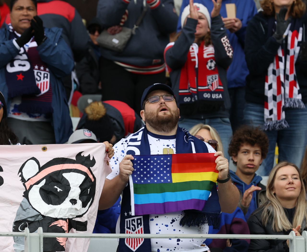 A bearded man in classes and a white speckled shirt holds a rainbow colored U.S. flag.