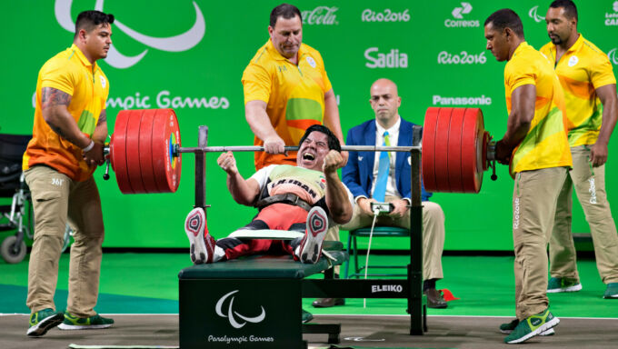 A paralympic weightlifter celebrates after bench pressing a bar with four large red plates on each side of a bar. Four men in yellow and orange polos stand as spotters.