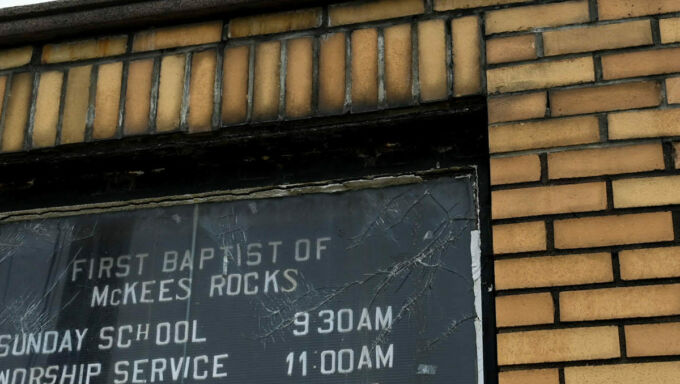 In front of a church steeple is signage on a black background ringed by tan bricks. The head of the sign reads First Baptist of McKees Rocks. Beneath that it reads Sunday School, 9:30AM, Worship Series, 11:00 AM, You are invited to join us. Featured text reads Pray for Damar Hamlin & Family.