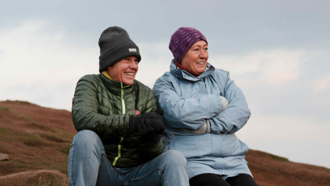 A couple sit on a rock near the top of a hill smiling and watching the sunrise. The person on the left is wearing a dark gray knit hat and green down jacket. The person on the right is wearing a pink and purple knit hat and a light blue rain jacket.