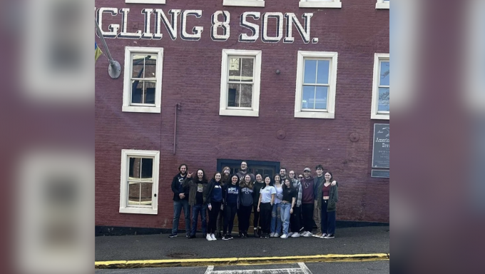 A group of students in the Penn State Brew Club stand outside the front entrance to a large, red brick building. The words "Yuengling and Son" Appear above them in white, stenciled lettering.