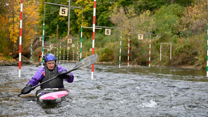 A man in a blue helmet, purple jacket, and black life vest propels a black and pink slalom kayak through a "gate" which consists of red and white horizontally-striped poles hanging above a creek.