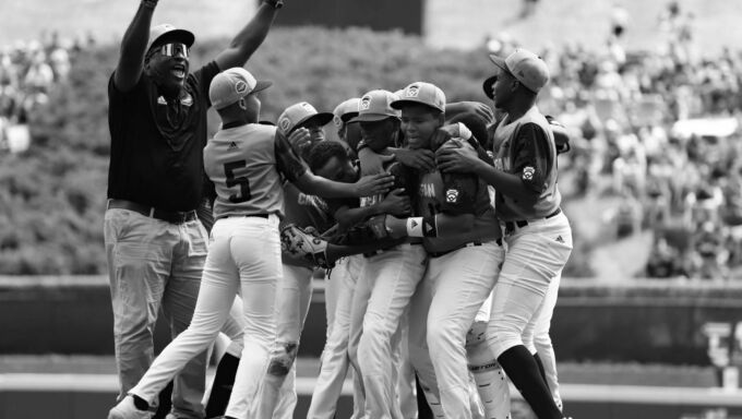 A team of little league baseball players hug each other and celebrate while their coach stands in the background with both arms raised excitedly in the air.