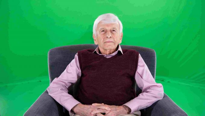 Gray haired older man in sweater vest seated in chair in front of green screen.