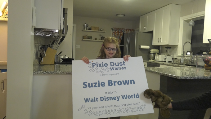 An image of Suzie Brown holding a sign that reads "Pixie Dust Wishes, Suzie Brown a trip to Walt Disney World" in a kitchen in State College