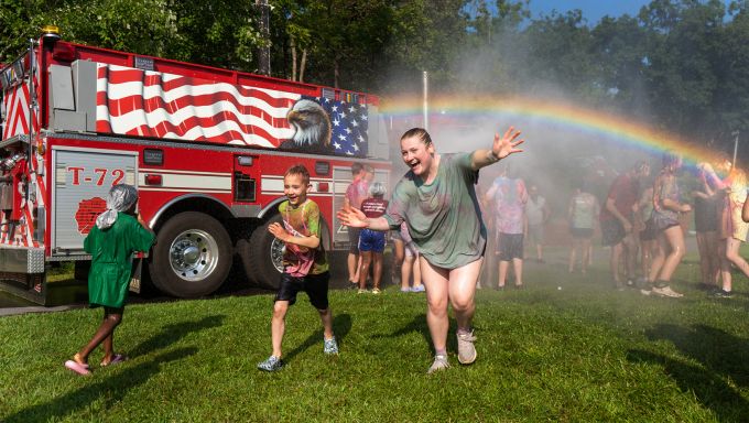 Children run in the rainbow spray of a water hose