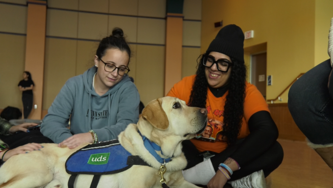 Two students sit behind a yellow lab wearing a "UDS" marked vest while petting him.