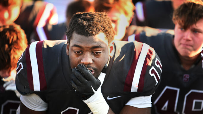 A football player in a gray, white, and maroon jersey holds a gloved hand over his mouth as tears roll down his face.