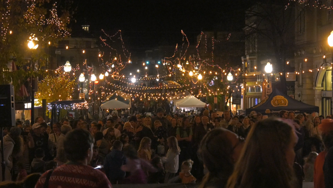 Lower Half: Hundreds of people fill Allen Street, Christmas market tents flank the sides of the road. Upper Half: Christms lights dangle across buildings on either side of Allen street.