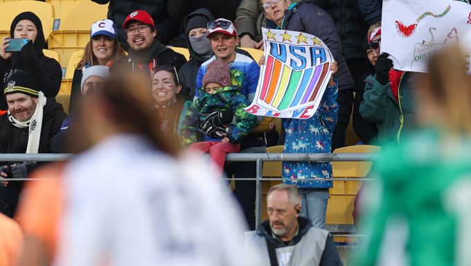 A young fan in a blue jacked with white unicorns holds a rainbow-colored USA Believe sign.