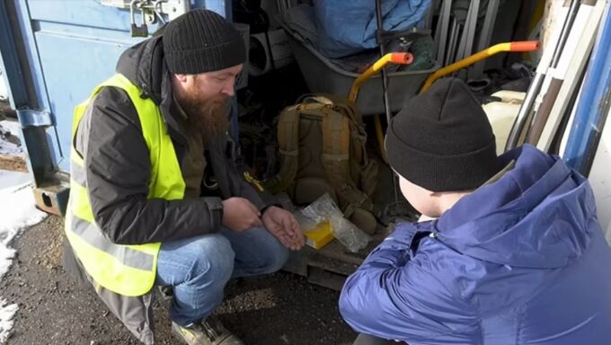 A man in a blue hooded jacket and black hat crouches and speaks with a man in a bright neon green safety vest and black beanie hat.