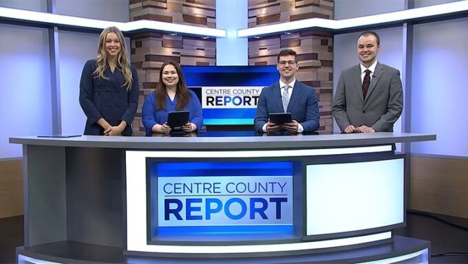 Four student news anchors sit and stand behind a newsdesk with a monitor that reads Centre County Report