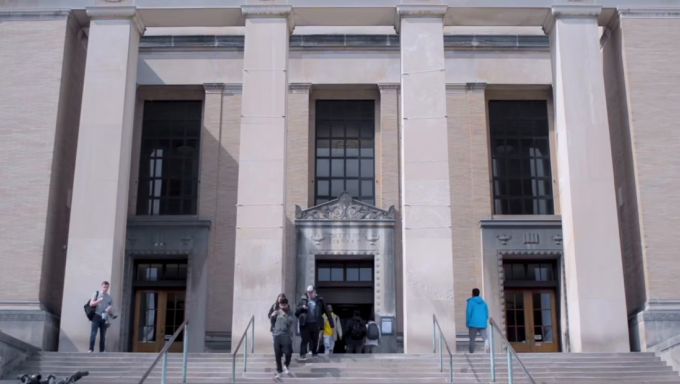 Students entering a large tan stone building with four pillars and windows gridded in black metal