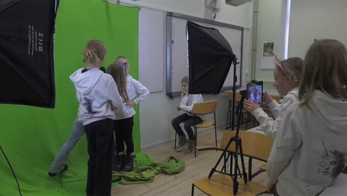 A crew of young girls in white hoody sweatshirts are filming a video in a studio in front of a green screen.