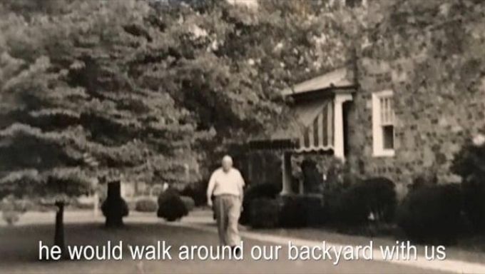 A man walks on grass in front of a house and a bunch of trees