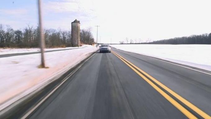 A car speeds down a two lane road with double yellow center line, snow on the roadside, with a silo in the background