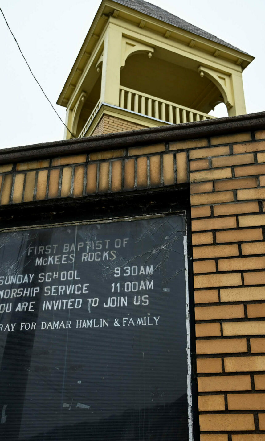 In front of a church steeple is signage on a black background ringed by tan bricks. The head of the sign reads First Baptist of McKees Rocks. Beneath that it reads Sunday School, 9:30AM, Worship Series, 11:00 AM, You are invited to join us. Featured text reads Pray for Damar Hamlin & Family.