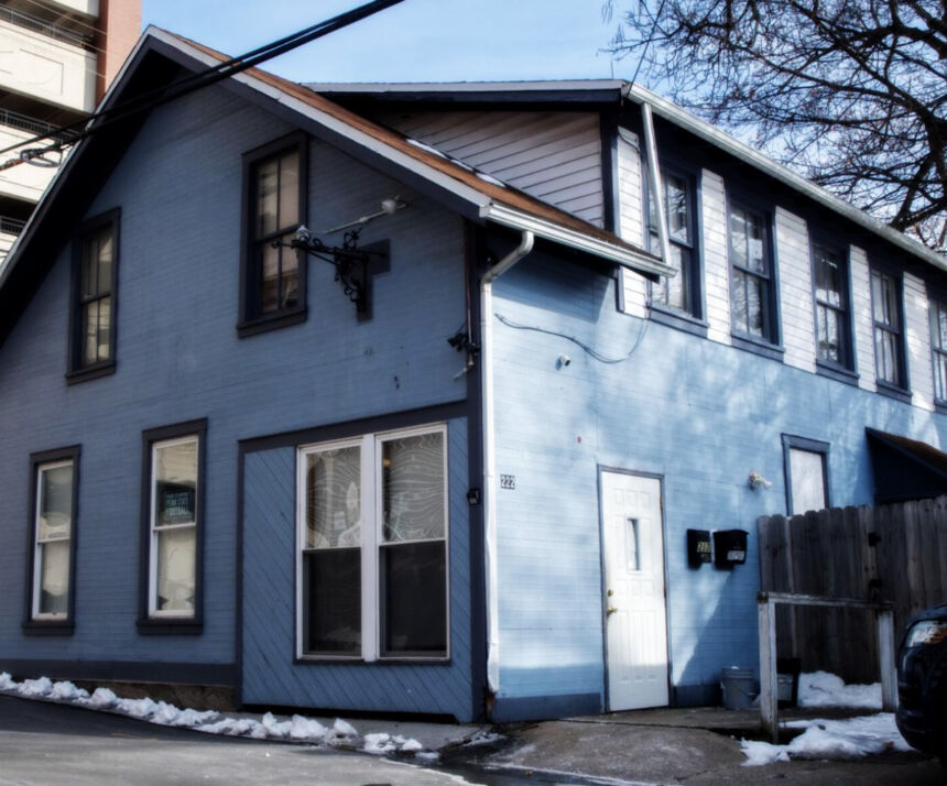 A blue two-story apartment house with small patches of snow on the ground in front. The side of the house near a sidewalk in the foreground has 5 windows. The side of the house has a white door and five windows on the second floor.