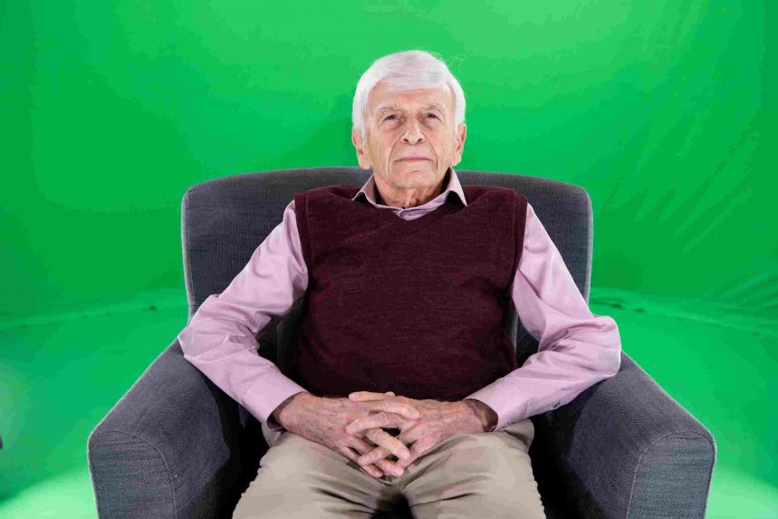 Gray haired older man in sweater vest seated in chair in front of green screen.