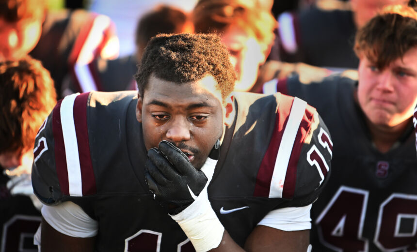 A football player in a gray, white, and maroon jersey holds a gloved hand over his mouth as tears roll down his face.