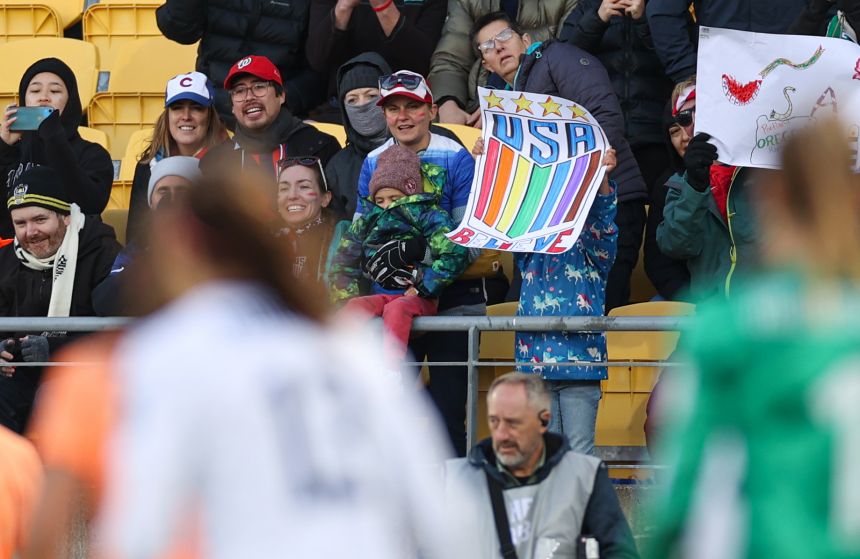 A young fan in a blue jacked with white unicorns holds a rainbow-colored USA Believe sign.